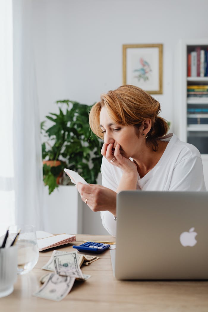 A woman reviews financial documents at a home desk with a laptop and cash visible.